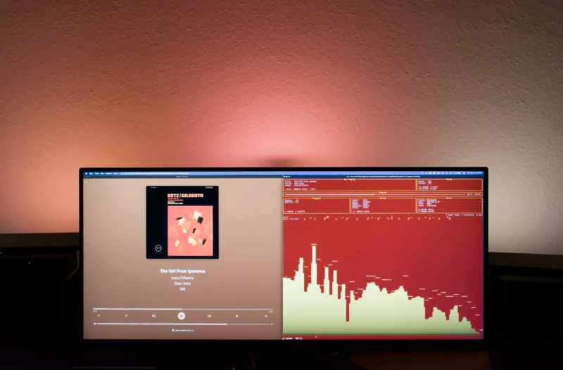 An ultrawide monitor in a dimly lit room displaying Music Assistant across two panels: the left showing the album Getz/Gilberto by Stan Getz playing 'The Girl from Ipanema,' and the right showing a green frequency visualizer on a red background. Warm orange ambient lighting glows behind the screen.